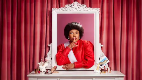 Shakespeare's Globe Anne Odeke wearing a crown and a red dress.
She is standing by a vanity table and has emerged herself in the mirror and has her index finger on her lips.