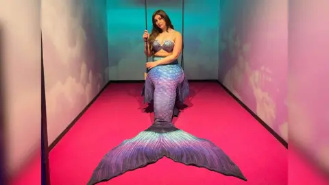 Jasmine Seales A woman sitting on a rope swing in a small room where the walls and floods are painted pink. She's wearing a shell bra and a large mermaid tail over her legs, in shades of iridescent blues, pinks, purples and blues.