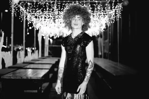 emma reed A candid photo of a drag queen smoking in an empty room