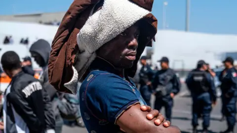 Getty Images A man part group of asylum seekers, mostly from African countries, waits for a negotiation commission to walk out El Chaparral port of entry during a protest in Tijuana, Baja California state on July 9, 2019, northwestern Mexico. - Asylum seekers claimed that Mexican migration officers were selling spots on the line to get an interview with US migration authorities.