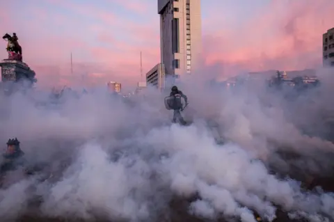 Eric Allende Gonzalez Police officer in riot gear runs amidst smoke bombs towards protesters