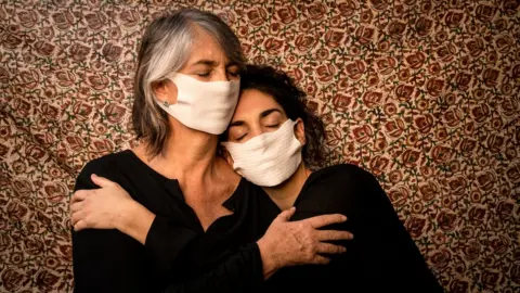 Tamara Merino/Covid Latam Two women embrace, wearing face masks