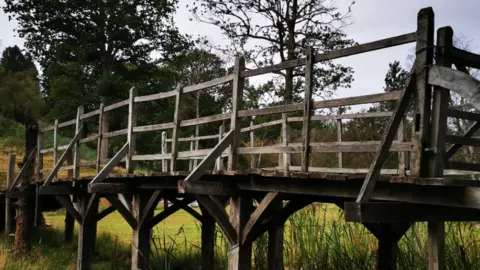 Summers Place Auctions Side angle view of rebuilt wooden bridge in a field over water