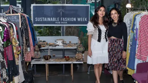 Sustainable Fashion Week India Priyanka Abhishek and Laghna Gowda standing near clothes rails by a Sustainable Fashion Week sign