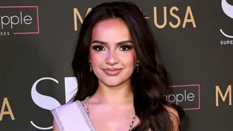 Getty Images Miss Teen USA 2023, UmaSofia Srivastava smiling at the camera, with a wall behind her which contains names of companies and Miss USA.