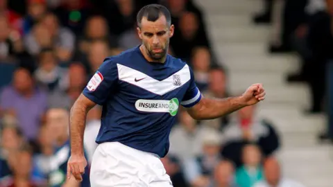 Southend United FC Chris Barker appears to be stretching to kick the ball during a match. He is wearing a blue Southend United strip and is wearing the captain's armband. There is a crowd sitting in the stands in the background.