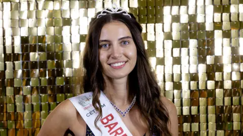 Miss England Head and shoulders image of Melisa Raouf wearing a tiara and sash that has the start of the word 'bare' on it. She is smiling and wearing no make up. She is standing in front of a gold, shimmering background