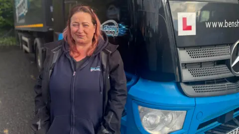 BBC Andrea stands in front of a truck and smiles at the camera. She has shoulder length pale red hair, wears a navy blue jumper and a black jacket.