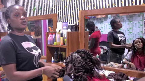 Ange Sea, wearing a black T-shirt with a female cartoon character in white printed on the front, adds extensions to a seated woman's hair in a salon in Daloa. The client looks into a wooden framed mirror.