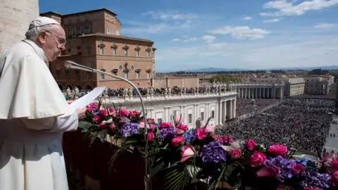 Reuters The Pope, on the left of the image, speaks into a microphone. He is standing on a balcony. Below, there is a large crowd of people. The sky is blue, with only a few clouds. In front of the Pope, there are flowers on the railing of the balcony. The Pope is dressed in white, ceremonial dress.