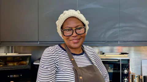 BBC A woman wearing a blue and white striped shirt, a brown apron and a white knitted hat smiles at the camera. She also wears black and gold glasses. She stands in a kitchen with grey cupboards and a microwave behind her.
