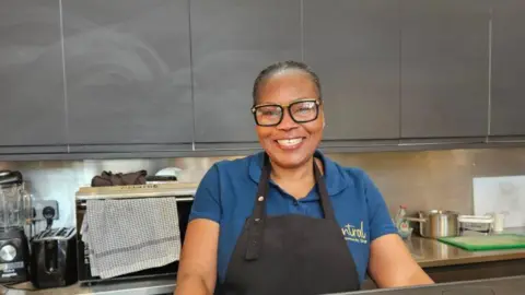 The Good Shepherd A woman in a navy blue shirt and black apron smiles at the camera. She stands in a kitchen, with silver cupboards behind her. On a stainless steel surface is kitchen utilities including a microwave and a blender.