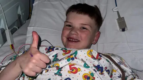 Máirtín Mac Gabhann Dáithí is lying in a hospital bed with tubes around him. He has short brown hair, smiling at the camera and is doing the thumbs up - he is wearing a patterned hospital gown with coloured numbers and cartoons.
