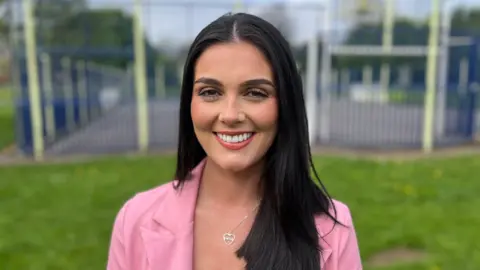 BBC A woman with long dark hair, wearing a pink jacket and a silver heart necklace, standing in a park with a basketball court in the background