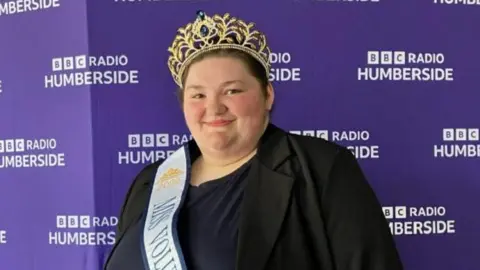 BBC Philippa Mackinder wearing a sash and the gold crown she won as Miss Voluptuous England. She is wearing a black and navy top and is standing in front of the purple BBC Radio Humberside backdrop.