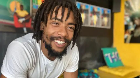 Jon Wright/BBC Donovan Charles Lansiquot sits in a community centre, smiling at the camera. There is bright artwork behind.