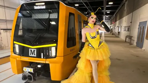 Nexus Sally TM posing to the right of one of the new dark yellow Metro trains. She has short, brown hair which is curled underneath, with a streak of yellow dye at the front. A piece of yellow hair is standing up above her head. She is wearing a yellow dress, which is lighter than the train, which is made up of a corset with silver accents and a tulle skirt which is short at the front and long at the back. She is also wearing a yellow collar around her neck and black long gloves.